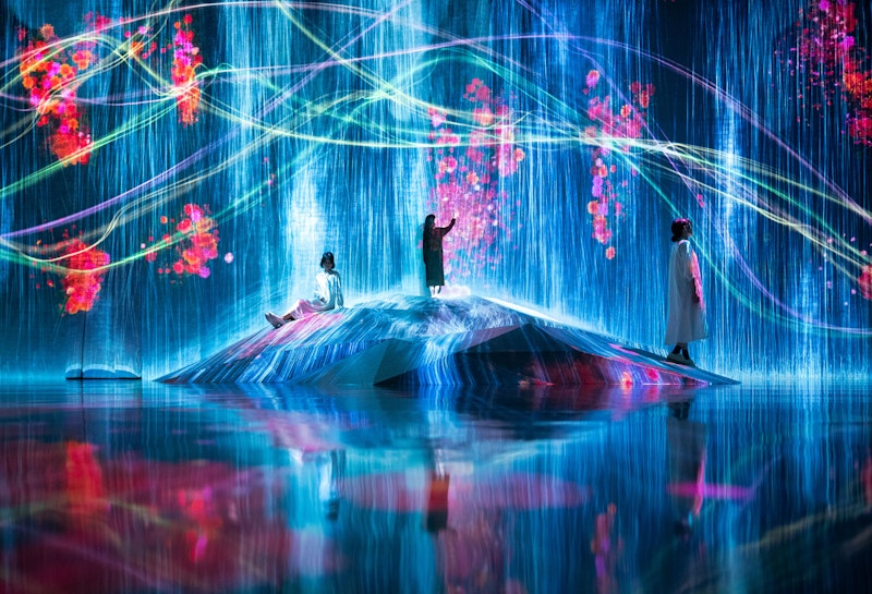 Three people interacting with an immersive digital art installation featuring flowing water particles and vibrant floral projections.